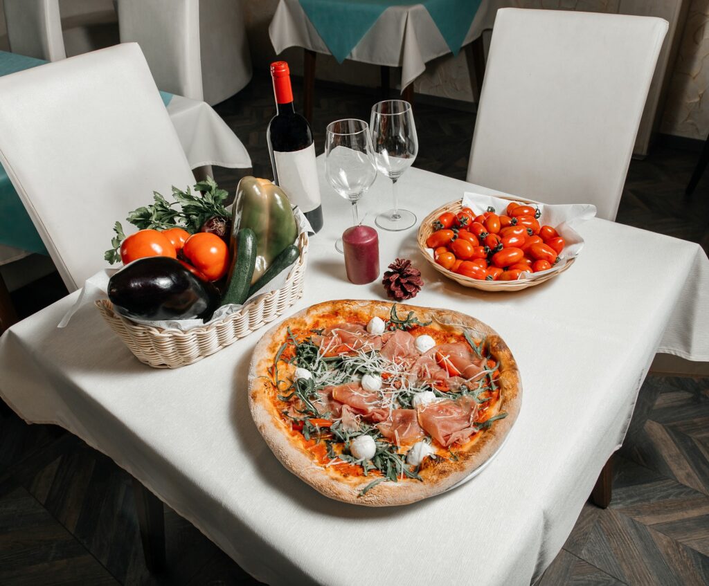 A pizza, a wine, two wine glasses, a basket of tomatoes, and a basket of vegetables on the table